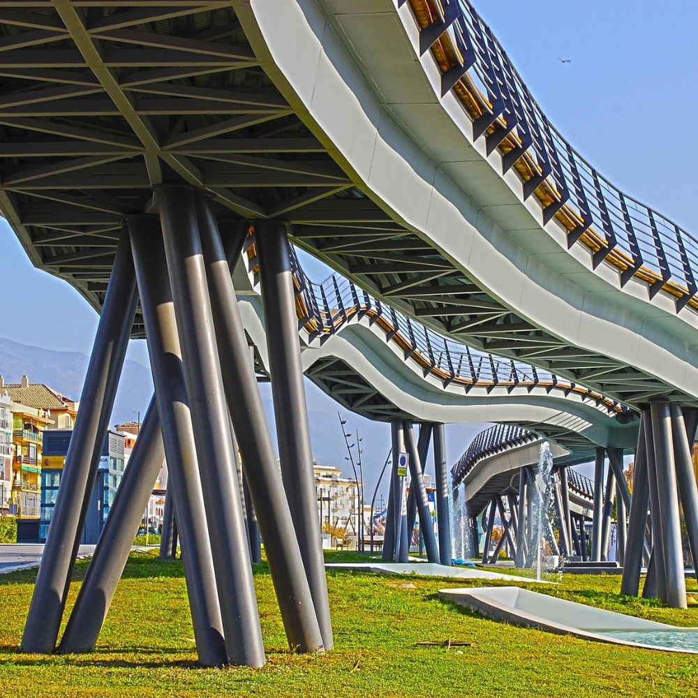 AbraCasaBra.se En modern gångbro med böljande, vågiga gångar som stöds av vinklade pelare sträcker sig över ett gräsbevuxet parkområde med vattenfontäner och byggnader som syns i bakgrunden.