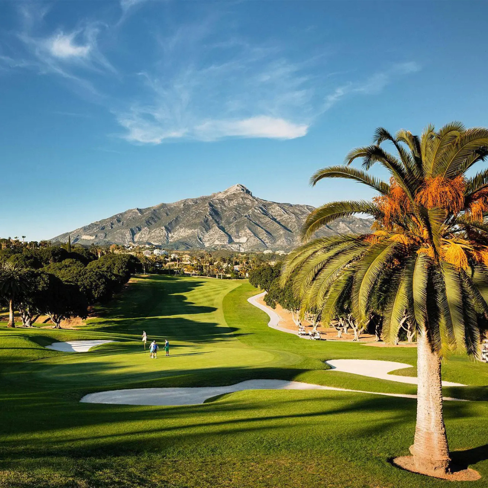 AbraCasaBra.se En naturskön golfbana med gröna fairways, sandbunkrar och höga palmer, med ett berg i bakgrunden under en klarblå himmel. Några golfare spelar på banan.