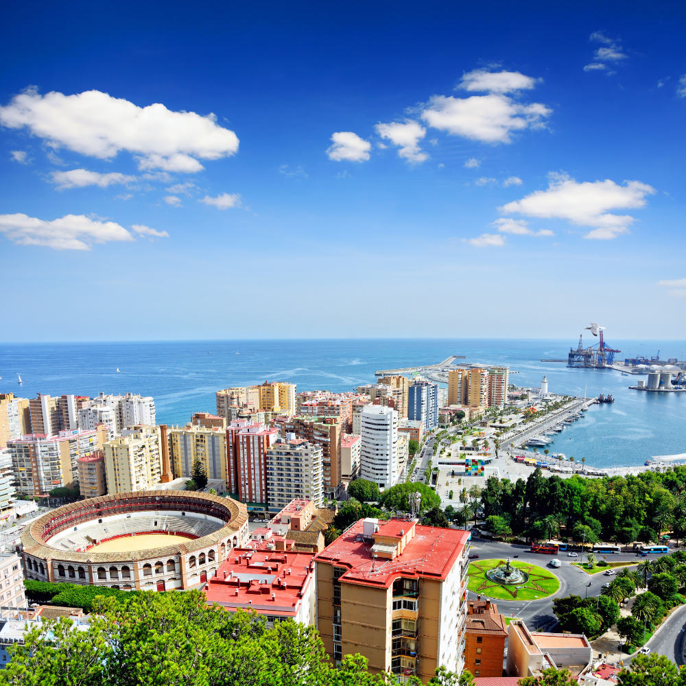 AbraCasaBra.se Flygfoto över en kuststad med en tjurfäktningsarena, moderna byggnader, en småbåtshamn och havet under en blå himmel med spridda moln.