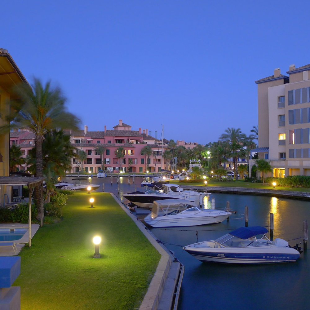 AbraCasaBra.se En fridfull marina i skymningen med båtar som ligger förtöjda längs en kanal, omgiven av moderna byggnader, palmer och mjuka trädgårdslampor som lyser upp den gröna gräsmattan och gångvägen.