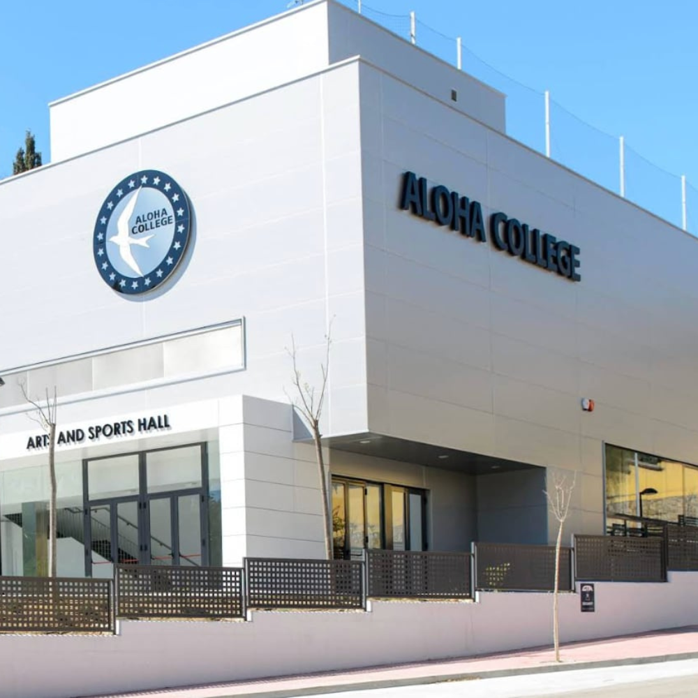 AbraCasaBra.se Modern vit byggnad med stora fönster och ALOHA COLLEGE-skylt på utsidan. På fasaden finns också Aloha Colleges logotyp och en skylt med texten ARENA AND SPORTS HALL. Svart stängsel löper längs framsidan.