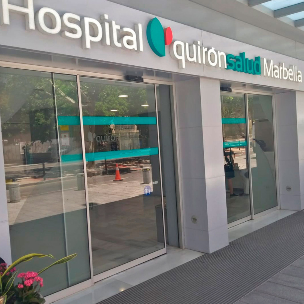 AbraCasaBra.se Glasentré till Hospital Quirónsalud Marbella, med sjukhusets namn ovanför dörrarna. På glaset syns reflexer och en person som tar ett foto. Träd och trafikkoner kan ses utanför.
