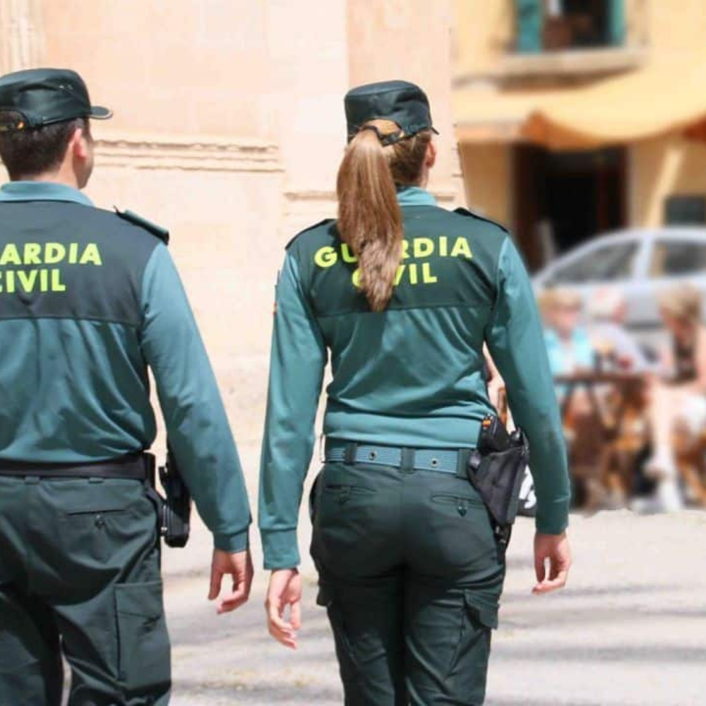 AbraCasaBra.se Två poliser från Guardia Civil, en man och en kvinna, går utomhus i uniform. Bilden är tagen bakifrån och bakgrunden är suddig med sittande människor och byggnader som syns.