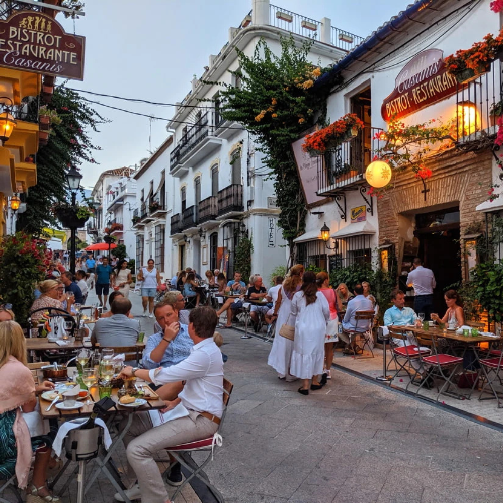 AbraCasaBra.se Uteservering på en charmig europeisk gata kantad av vita byggnader och balkonger; människor njuter av mat och dryck vid borden när kvällen faller på, under varma lampor och blomsterdekorationer utanför en bistrorestaurang.