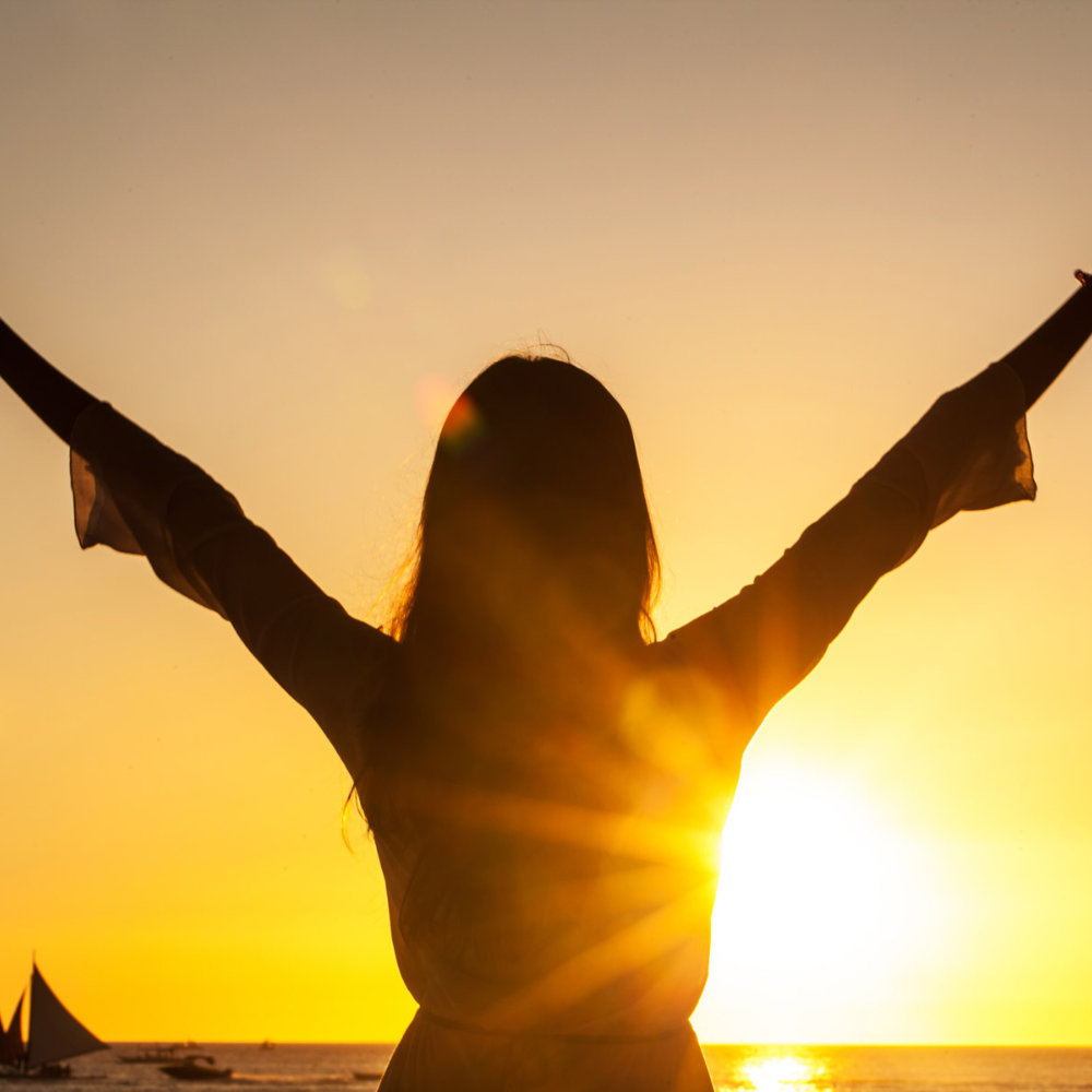 AbraCasaBra.se En person står med armarna i vädret i siluett mot en ljus solnedgång över havet. En segelbåt syns på vattnet och himlen lyser i orange och gult.