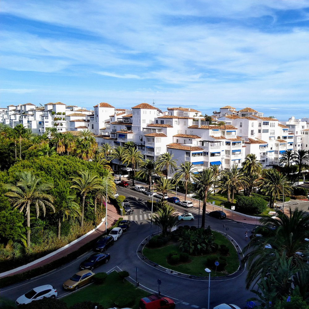 AbraCasaBra.se En rondell med palmer och parkerade bilar ligger framför vita hyreshus i medelhavsstil under en blå himmel nära kusten.