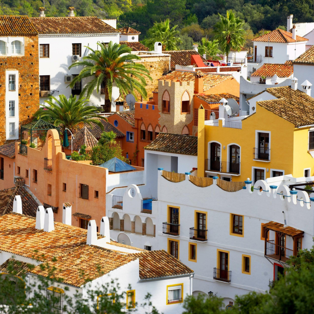 AbraCasaBra.se Färgglada byggnader i medelhavsstil med terrakottatak, palmer och balkonger, tätt packade på en sluttning, vilket skapar en livlig och pittoresk bybild.
