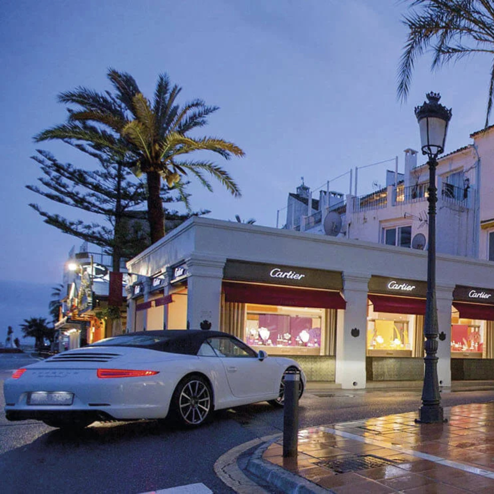 AbraCasaBra.se En vit lyxig sportbil svänger runt ett hörn nära en Cartier-butik med ljusa skyltfönster, palmer och våt trottoar i skymningen.