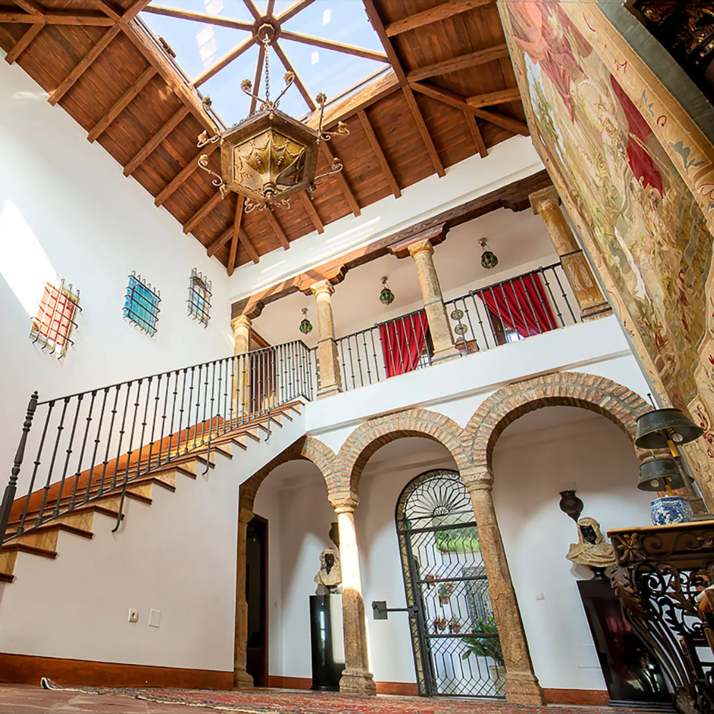 AbraCasaBra.se Ljust atrium med takbjälkar i trä, takfönster, utsmyckad ljuskrona, stenpelare, välvda dörröppningar, trappräcke i smidesjärn, gobelänger och statyer som blandar traditionella och historiska arkitektoniska stilar.